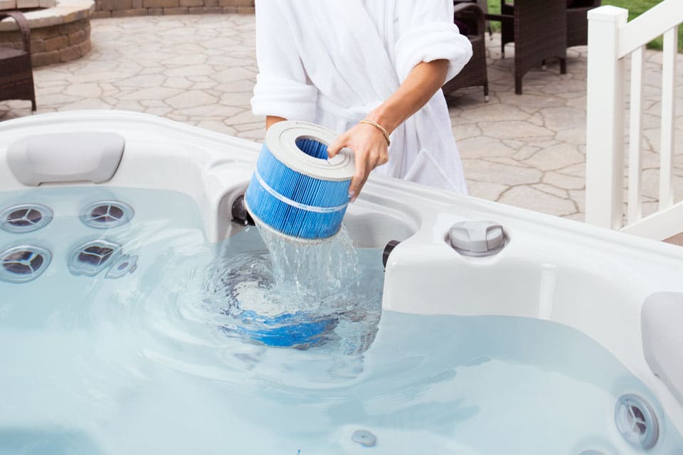 A person is shown replacing a hot tub filter. They are wearing a robe and standing next to the hot tub in a backyard.